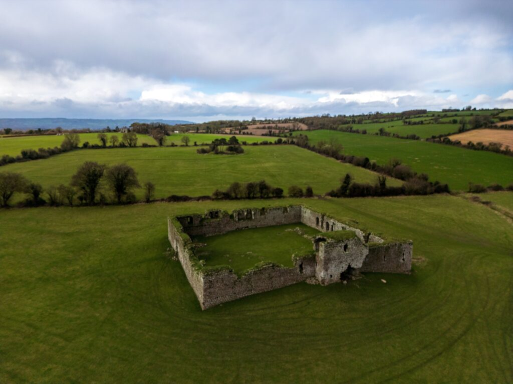 An aerial view of Ballymoon Castle near Muine Bheag (Bagenalstown) in County Carlow, Ireland. The ruin stands alone in a wide grazed field: a roofless square enclosure of thick granite walls, moss-streaked and broken along the top, with a t