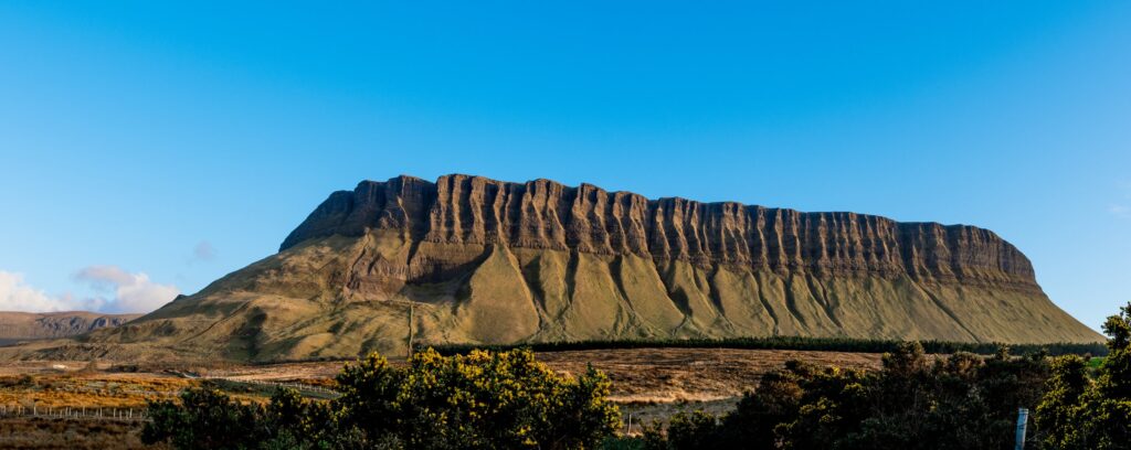 Benbulbin in County Sligo, Ireland, the flat-topped limestone mountain often called Ireland's Table Mountain, photographed in wide panorama under a cloudless deep blue sky. Low golden sunlight rakes across its steep fluted flanks, picking o