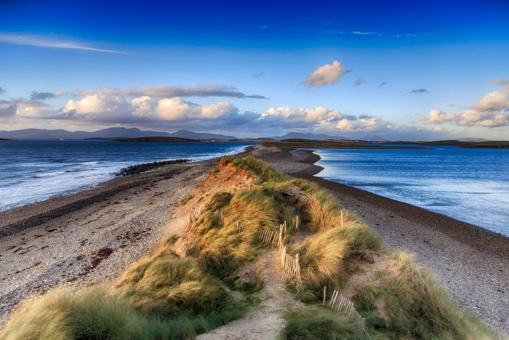 Sandy spit with marram grass dunes extending into blue waters under cloudy sky