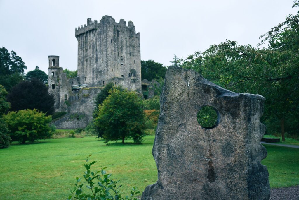 Blarney Castle in County Cork, Ireland, its tall grey stone keep with a crenellated and machicolated parapet rising above trees on a low bluff, with a ruined round tower to the left. A large upright standing stone with a circular hole bored