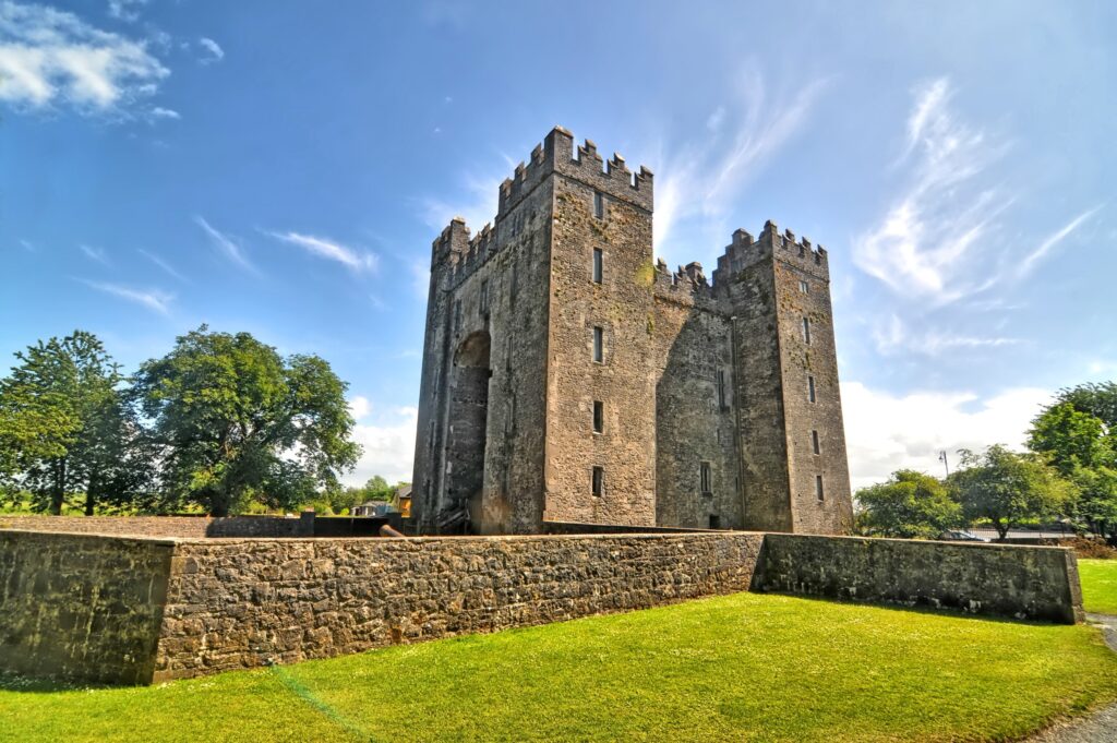 Bunratty Castle in County Clare, Ireland, photographed from below across a mown grass bank and a stone revetment wall on a bright summer day. The grey limestone tower house rises as a massive rectangular block with four square corner towers