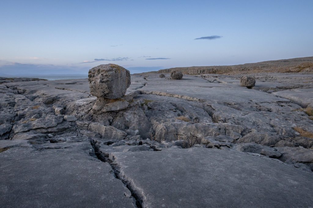 Bare limestone pavement in the Burren National Park, County Clare, Ireland, at dusk. Deep grikes and fissures split the flat grey clints, and several rounded glacial erratic boulders sit perched on the bedrock, the nearest resting on a smal