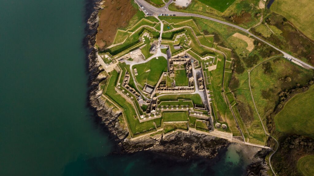 A near-vertical aerial view of Charles Fort at Summercove near Kinsale, County Cork, Ireland, in a wide letterbox crop. The star-shaped fortification occupies the centre of the frame on a green headland: five angled bastions of grey stone w