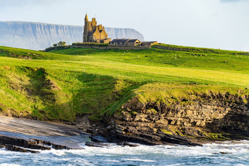 Classiebawn Castle on Mullaghmore Head, County Sligo, Ireland, in low evening sunlight. The turreted sandstone house stands alone on a green headland above layered limestone sea cliffs, with sheep grazing the fields and fence lines running