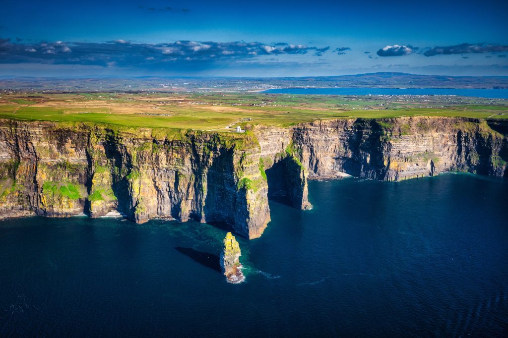 An aerial view along the Cliffs of Moher in County Clare, Ireland. The layered sandstone and shale cliffs run away in a series of dark headlands, their faces streaked with bright green where grass clings to the ledges, and the free-standing