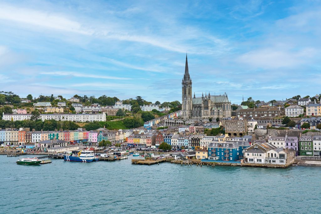 Elevated view across Cork Harbour to the waterfront of Cobh, with rows of brightly painted pastel terraced houses climbing the hillside and the tall Gothic Revival spire of St Colman's Cathedral rising above the town, a green ferry and a bl