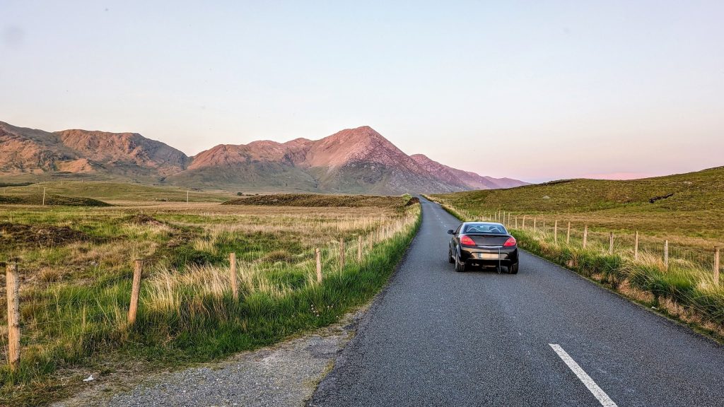 A single dark hatchback drives away from the camera along an empty two-lane road running dead straight through open bog and rough pasture in Connemara, County Galway. Post-and-wire fencing lines both verges, the grass is pale winter straw o