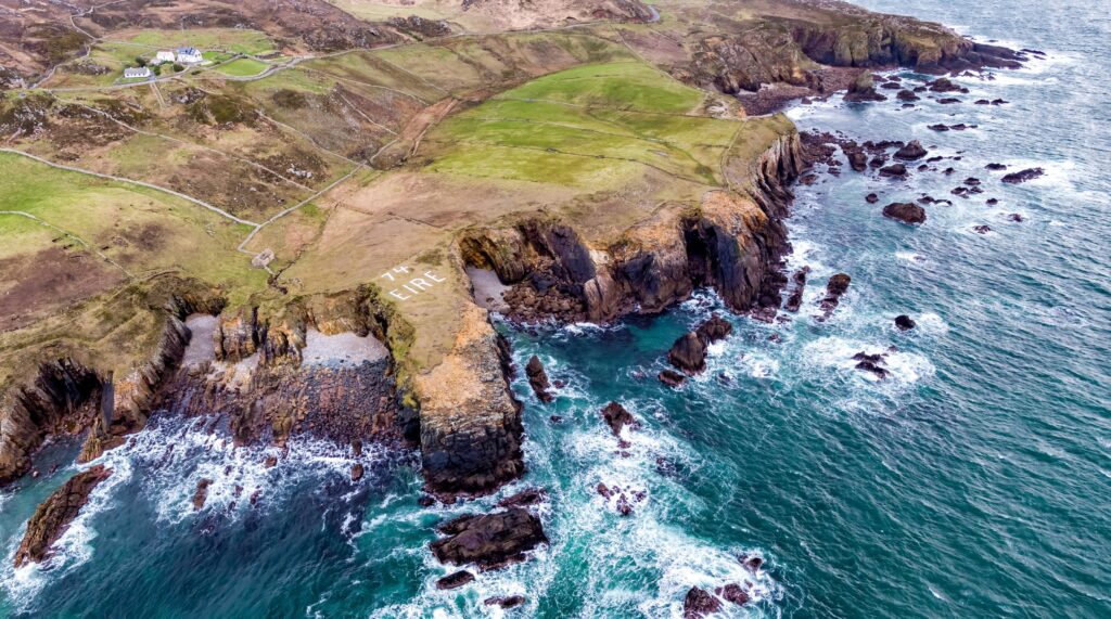 An aerial view of the Atlantic coast near Maghery and Dungloe in County Donegal, Ireland, looking down on layered sea cliffs, stacks and reefs with white surf breaking over them in deep teal water. On the flat clifftop, picked out in white