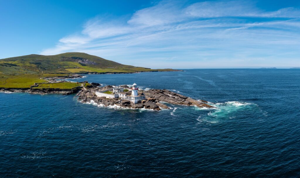 A drone view of the lighthouse at Cromwell Point on Valentia Island, County Kerry, Ireland. The short white tower with its lantern stands inside a low white-walled compound of keepers' buildings on a spit of dark rock reaching out into the