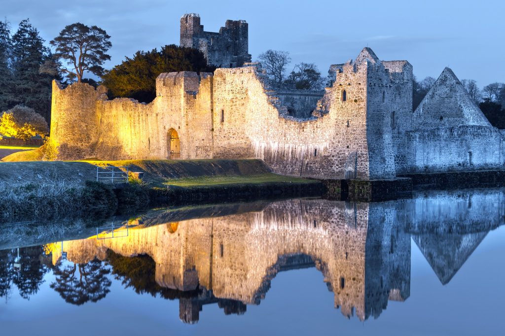 Desmond Castle at Adare in County Limerick, Ireland, floodlit at blue hour and mirrored almost perfectly in the still water of the River Maigue. The ruin runs right across the frame: a low curtain wall with an arched gateway and a wooden do