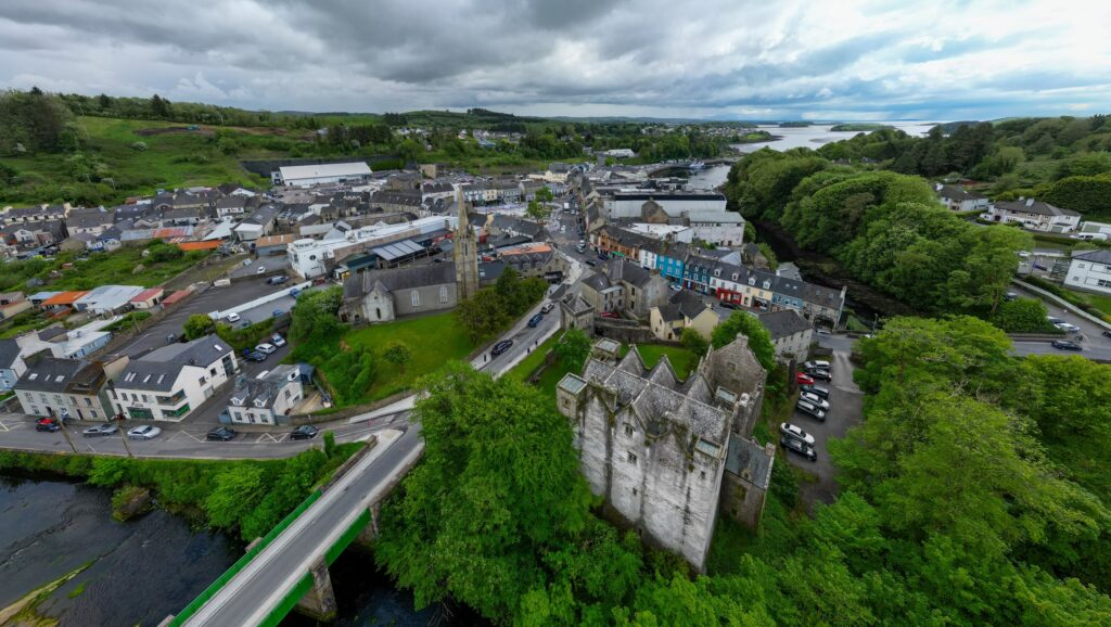 A wide panoramic drone view over Donegal Town on 14 May 2024, taken from above the River Eske looking north across the settlement. Donegal Castle stands in the right foreground among heavy fresh green trees, its grey tower house and gabled