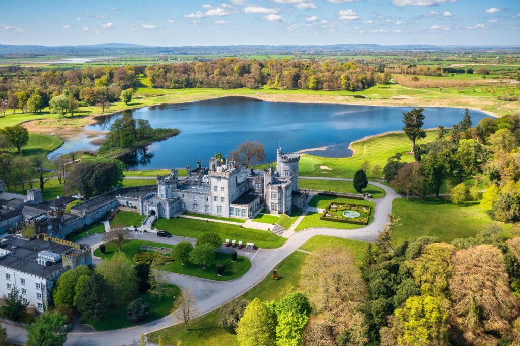 An aerial view of Dromoland Castle near Newmarket-on-Fergus, County Clare, Ireland, on a bright spring or early summer day. The grey limestone castle stands centre left: a long battlemented range with corner turrets, a round tower, tall chi