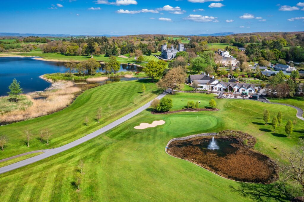 An aerial view over the golf course at Dromoland Castle, County Clare, Ireland. The foreground and middle of the frame are given over to the course: broad mown fairways in stripes of light and dark green, a putting green with a flag, a pale