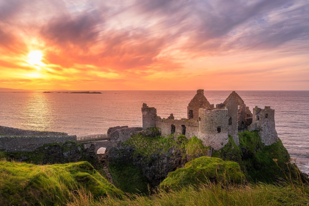 The ruins of Dunluce Castle on the North Antrim coast near Bushmills, Northern Ireland, standing on a grassy basalt headland above the Atlantic at sunset. The roofless gables, round towers and curtain walls are lit warm by low sun, with a n