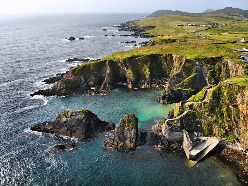 An aerial view of Dunquin Pier on the Dingle Peninsula, County Kerry, Ireland. A narrow concrete ramp switchbacks down the face of a green-topped sea cliff to a small concrete jetty at the water's edge, with dark layered sea stacks rising o