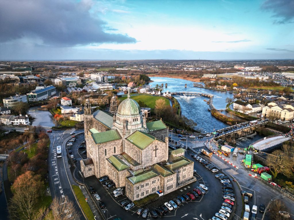 An aerial view over Galway Cathedral on Nun's Island, with the River Corrib winding away north through the city towards open countryside and a bank of dark cloud clearing to blue. The cathedral's pale limestone bulk fills the lower centre o