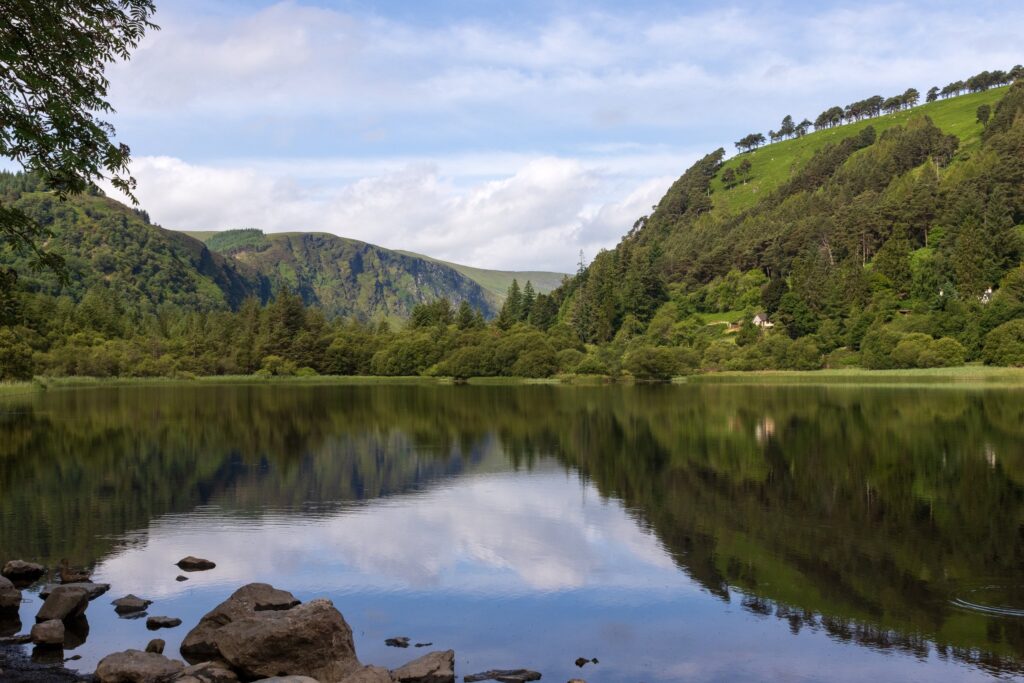 The Lower Lake at Glendalough in the Wicklow Mountains, County Wicklow, Ireland. The lake fills the lower half of the frame, almost completely still and holding a clean mirror image of the wooded slopes and cloud above; a scatter of grey bo