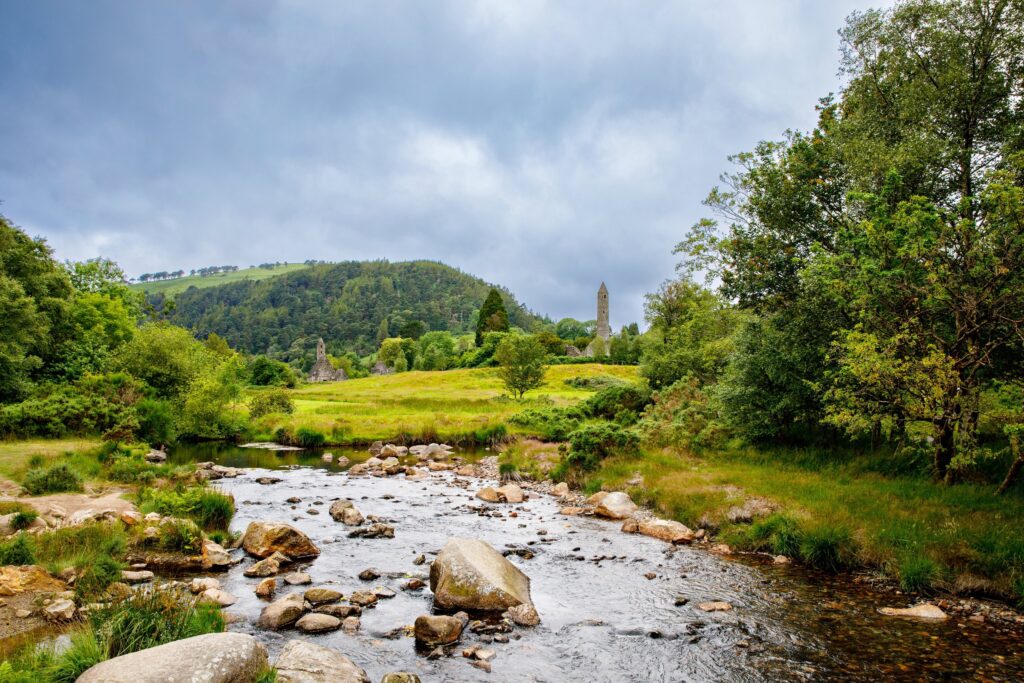 A shallow boulder-strewn river runs through the Glendalough valley in County Wicklow, Ireland, with the round tower of the early monastic settlement standing above the trees in the middle distance. Large pale granite boulders and cobbles fi
