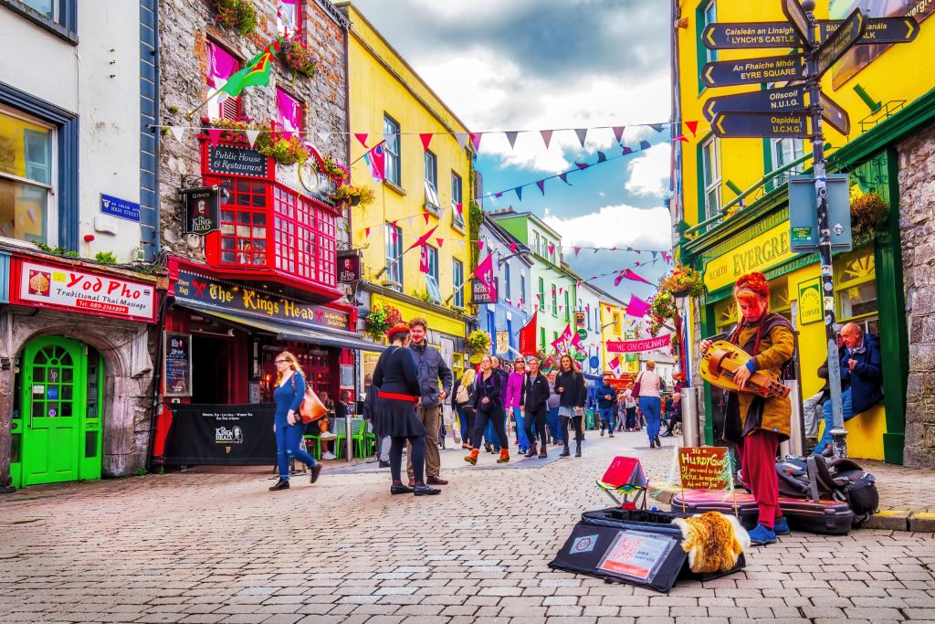 Colorful storefronts line a cobblestone street with pedestrians and street musicians in Galway