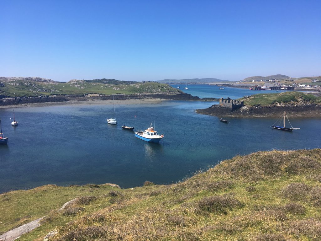The harbour of Inishbofin, an island off the Connemara coast of County Galway, Ireland, seen from a grassy headland on a clear summer day. Calm blue water fills the middle of the frame with a small blue-and-white fishing boat, sailing yacht
