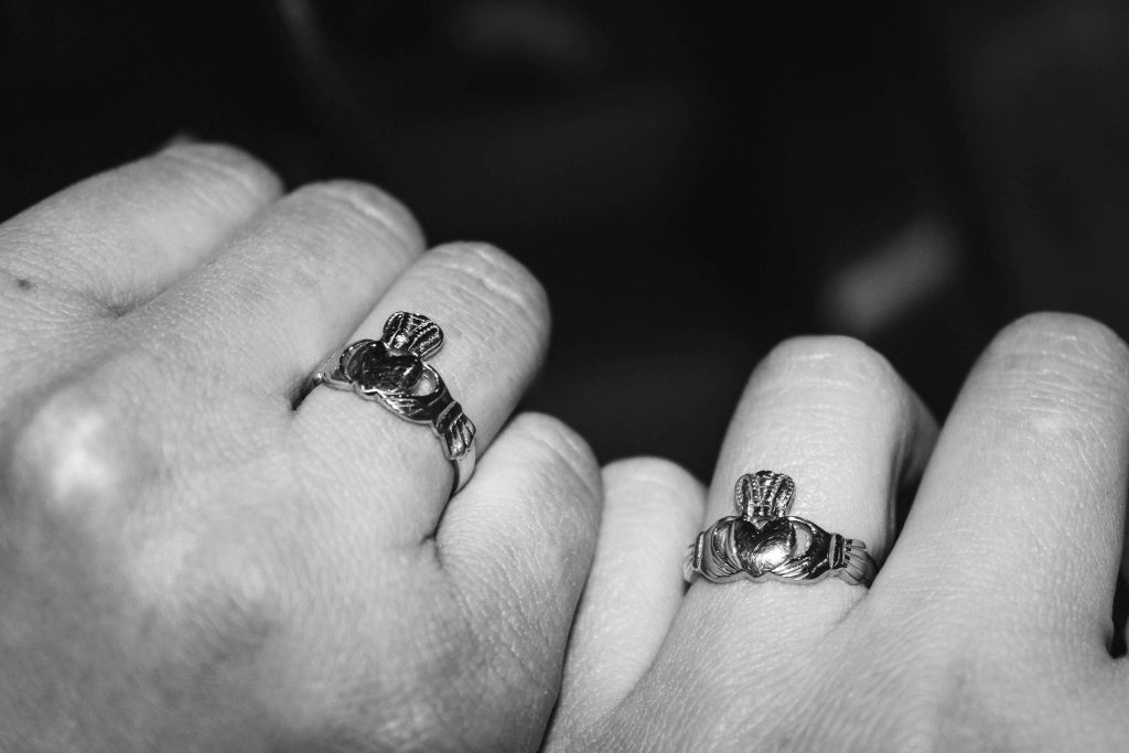 A close black-and-white still life of two hands resting side by side against a dark background, each wearing a silver Claddagh ring on the ring finger - the traditional Irish design of two hands clasping a crowned heart, standing for love,