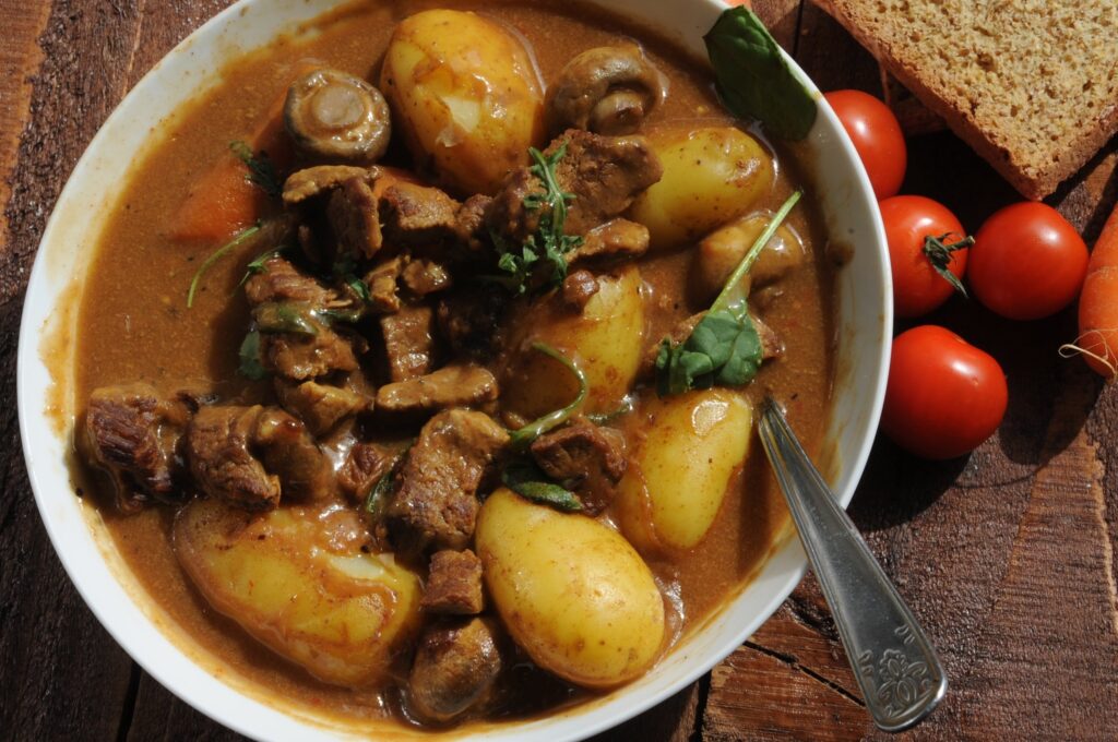 A white bowl of beef stew photographed close and from above on a rough dark wooden table. Chunks of braised beef, whole small new potatoes with the skins on, halved mushrooms and a slice of carrot sit in a thick brown gravy, scattered with