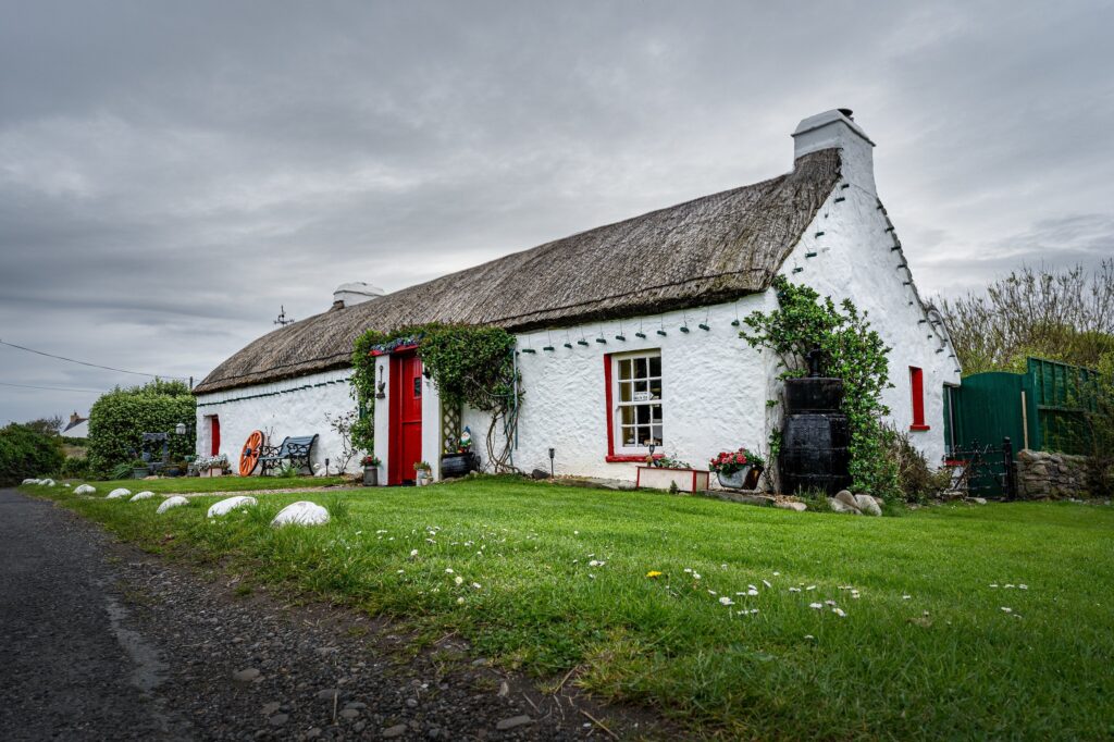 A traditional Irish thatched cottage, whitewashed and single-storey, photographed from the gravel lane that runs past its gable end under a heavy grey sky. The long low building runs diagonally across the frame from the left: rendered walls