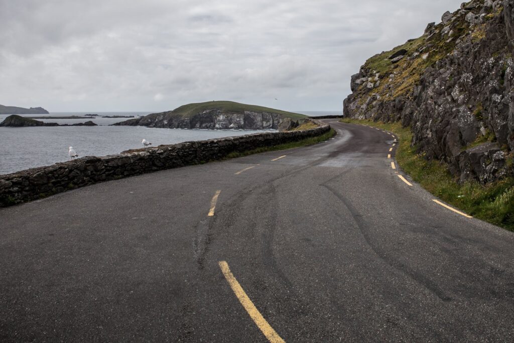 A narrow coastal road curving along the edge of a headland in western Ireland on an overcast day. The tarmac fills the lower half of the frame with worn yellow edge lines and a broken centre line; a low dry stone wall runs along the seaward