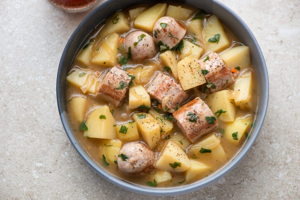 A high-angle shot of a grey-blue ceramic bowl of sausage and potato stew, filled almost to the rim and set slightly left of centre on a pale beige speckled stone surface. Large irregular chunks of peeled potato sit in a thin golden broth wi