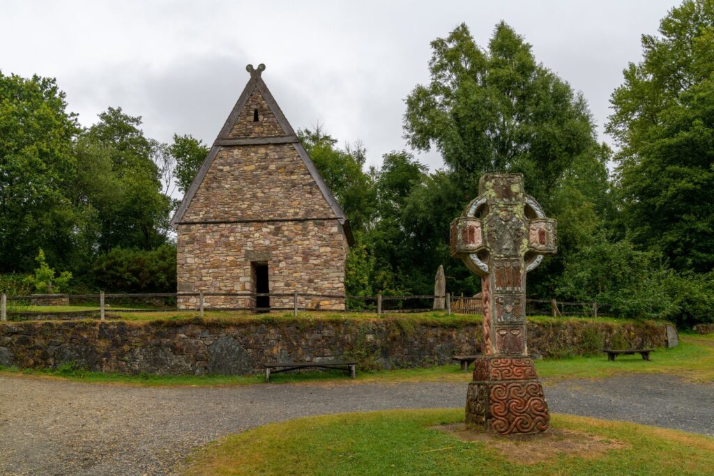 A reconstructed early Christian monastic enclosure at the Irish National Heritage Park near Wexford, photographed on 18 August 2022. In the centre foreground stands a tall high cross carved with interlaced Celtic ornament and inset panels,