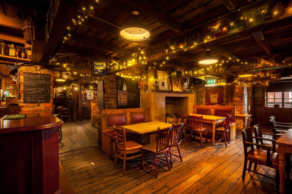 The empty interior of a traditional Irish pub in Kildare town, County Kildare, lit entirely by warm yellow lamps and strings of fairy lights looped across heavy dark timber ceiling beams. Bare brick and plaster walls, a curved dark wood bar