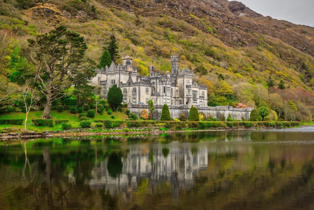 Kylemore Abbey in Connemara, County Galway, Ireland, seen across the still water of Pollacapall Lough. The long grey granite house stands on the north shore at the foot of a steep mountainside: battlemented parapets, a turreted tower, tall
