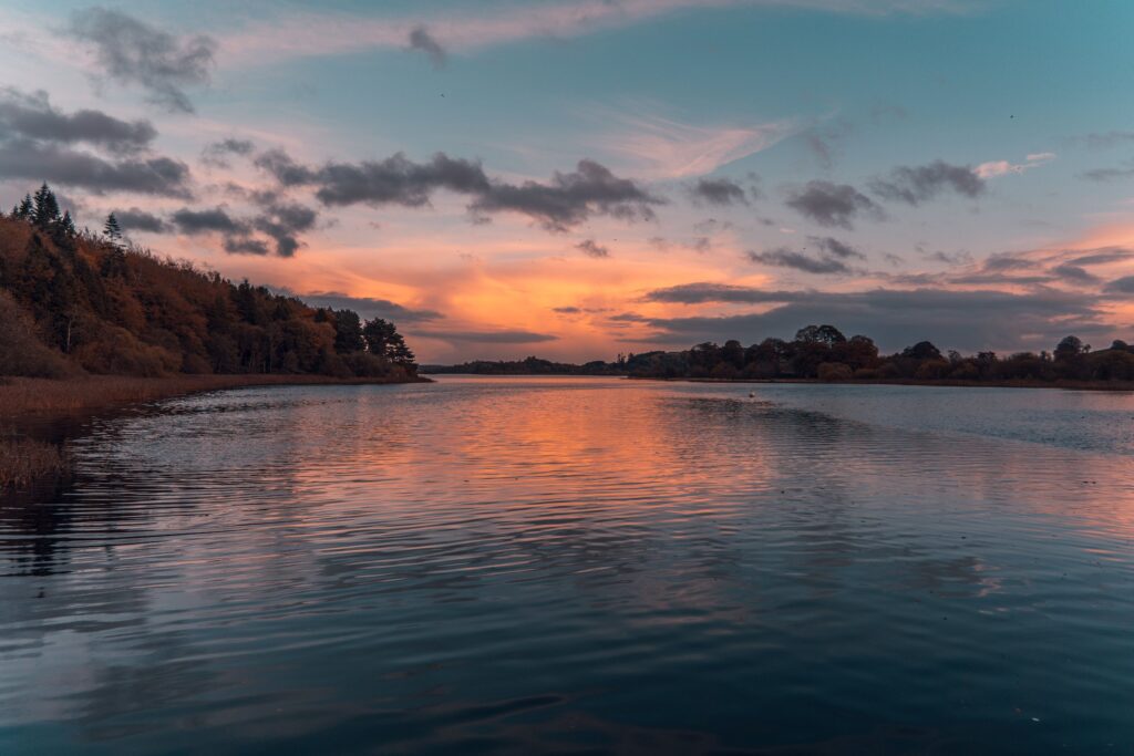 Sunset over Lough Muckno at Castleblayney in County Monaghan, Ireland, looking down the lake from a low vantage near the shore. The sky is banded in orange, salmon and pale gold behind a broken layer of grey-violet cloud, with the brightest