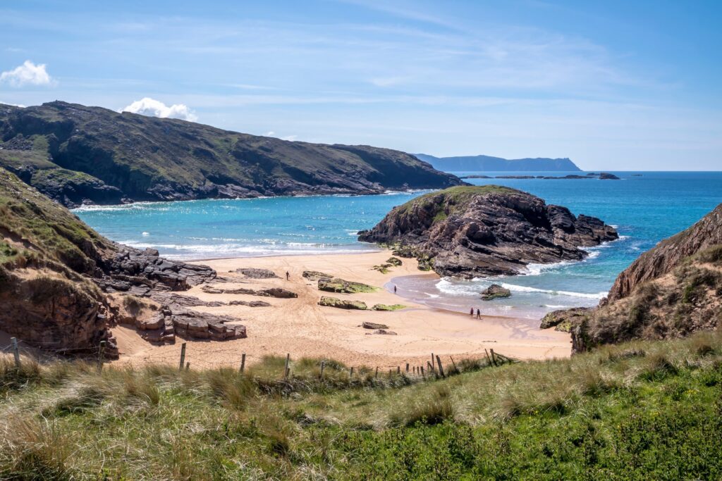Murder Hole beach, officially Boyeeghter Bay, on the Rosguill peninsula in County Donegal, Ireland, seen from the grassy dunes above. A curve of pale golden sand runs between low rocky headlands with a large rounded rock outcrop in the midd