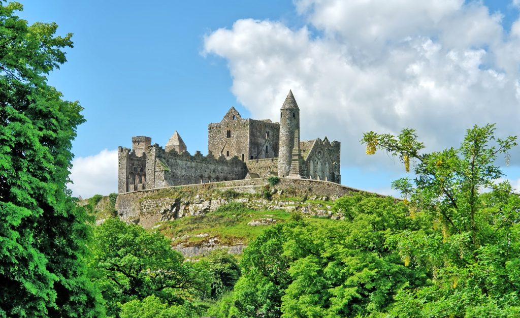The Rock of Cashel in County Tipperary, Ireland, seen from below across a steep green slope of trees and scrub on a bright summer day. The grey limestone group stands along the crest of the limestone outcrop: the roofless Gothic cathedral w