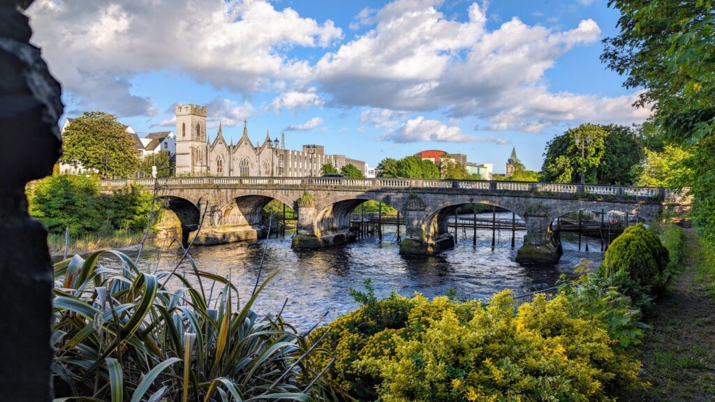 The Salmon Weir Bridge over the River Corrib in Galway city, Ireland, in a wide letterbox crop. The grey limestone bridge crosses the middle of the frame on a series of low arches, its parapet and lamp standards catching the sun, with the w