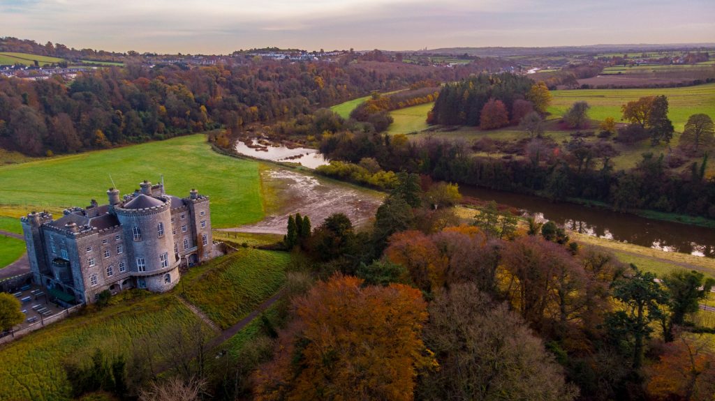 An aerial view of Slane Castle in its demesne above the River Boyne, County Meath, Ireland, in late autumn light. The grey castellated house sits on a bluff at the left with lawns falling away below it; the Boyne loops through the middle di