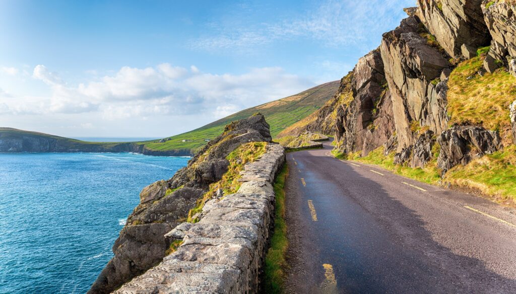 The Slea Head Drive on the Dingle Peninsula, County Kerry, Ireland, seen from the roadside looking west. The single-carriageway road with a broken yellow edge line curves in from the bottom right and disappears round a rock buttress in the