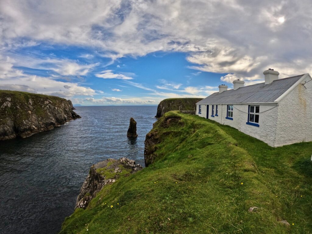 A long single-storey whitewashed cottage stands on the grassy lip of a sea cliff in County Donegal, Ireland, its gable and side wall filling the right of the frame. The walls are rough-cast and limewashed white, the window reveals and frame