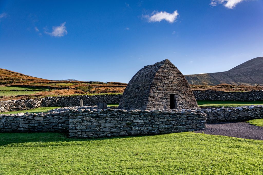 The Gallarus Oratory, an early Christian dry-stone church shaped like an upturned boat, stands within a low dry-stone enclosure wall on the Dingle Peninsula, County Kerry. Its corbelled grey stone walls and small dark doorway catch bright w