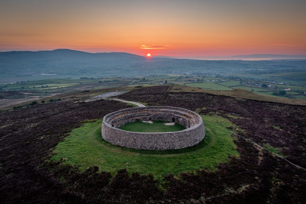 An aerial view of the Grianan of Aileach, the ancient ringfort on Greenan Mountain in County Donegal, Ireland, at sunrise. The circular drystone cashel sits centrally on the hilltop, its thick battered wall of grey rubble enclosing a grasse