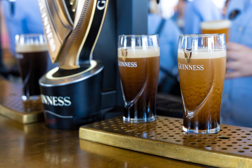 Two freshly poured pints of Guinness settling on a brass drip tray beside a black and gold harp-shaped Guinness tap, the stout's surge still visible as the creamy heads form, with a third dark pint behind and blurred visitors holding drinks