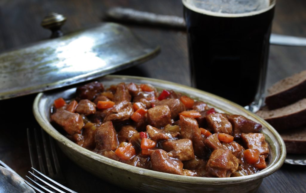A beef and stout stew served in an oval dish on a dark wooden table, styled for St Patrick's Day. The dish sits low in the frame, a shallow oval in dull brass or pewter with a wide rim, filled with chunks of braised beef in a glossy dark gr