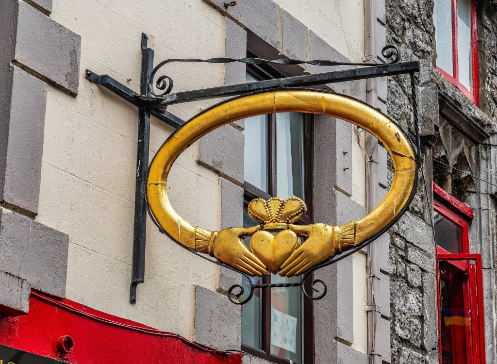 A large gilded Claddagh ring hanging as a shop sign from the wall of a stone building, photographed close up from below. The sign is a three-dimensional carved and gilded replica of the traditional Irish ring: a thick oval band in bright go