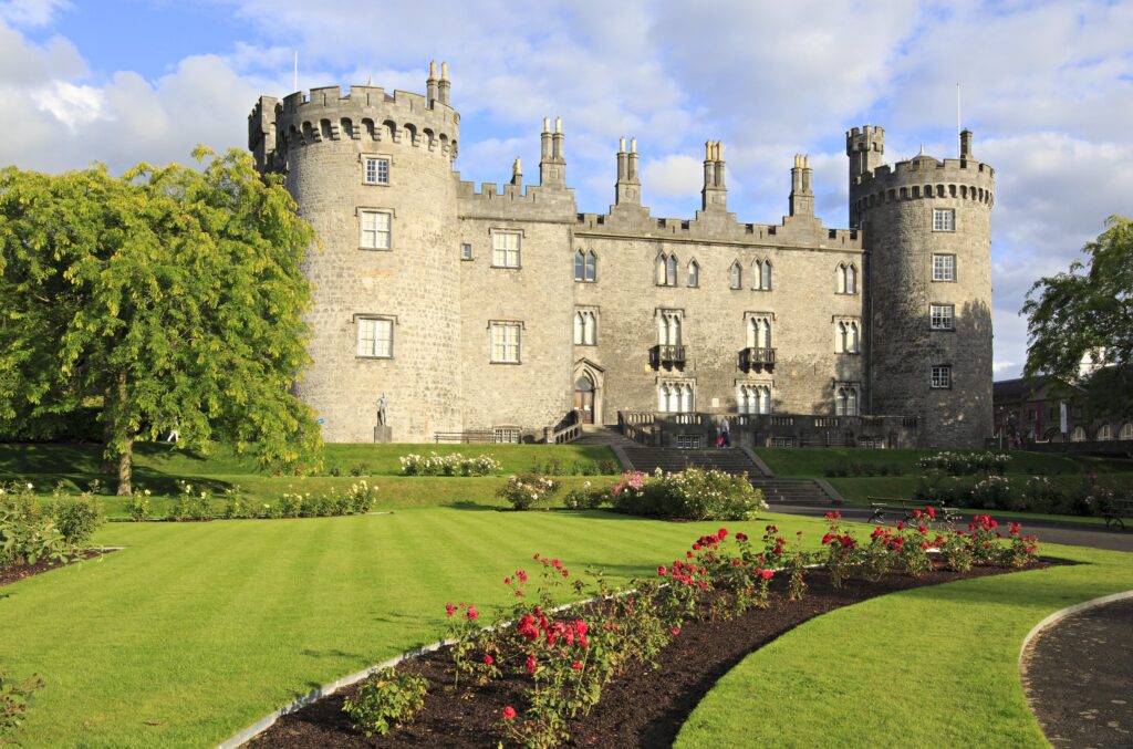 Kilkenny Castle in warm evening sunlight, seen from its formal gardens: the grey limestone castle with two round crenellated corner towers, tall chimneys and rows of Gothic-style windows rises above a broad stone staircase and terrace. Stri