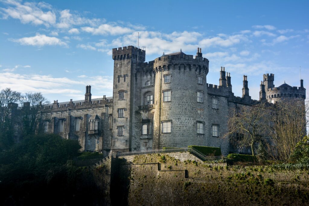 Kilkenny Castle seen from the river side on a clear winter day, rising above its terraced retaining walls. The grey limestone building fills the frame at an angle: a big round drum tower on the right with a machicolated parapet and tall chi