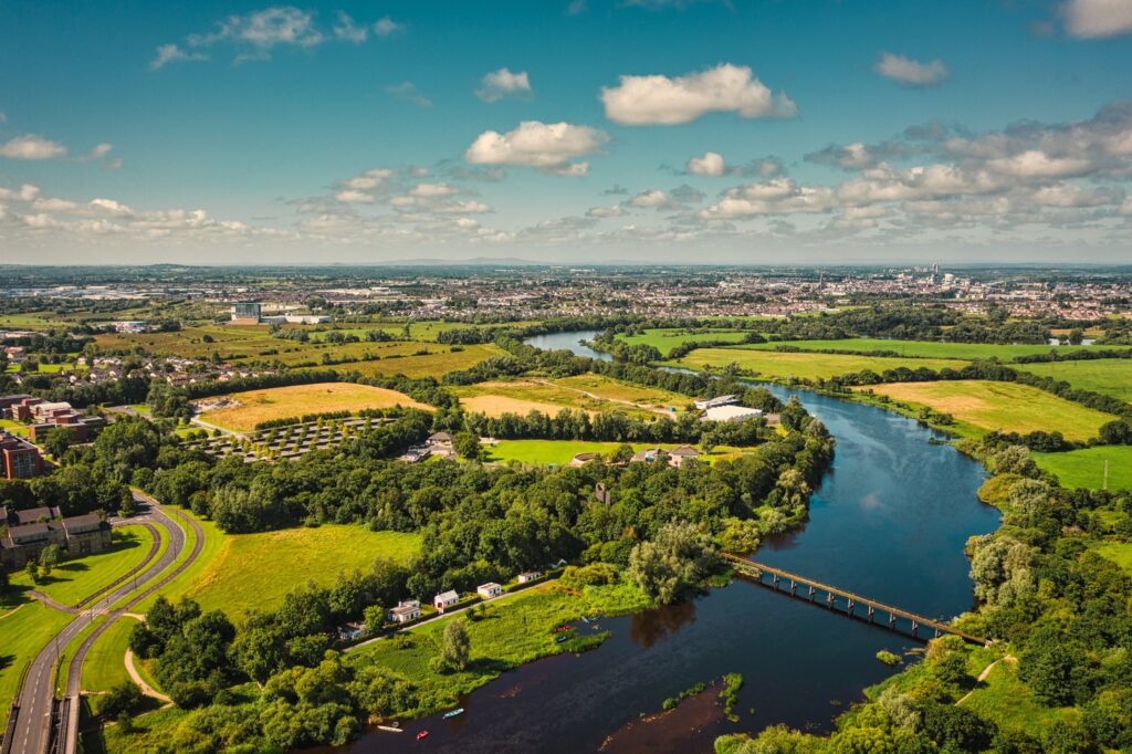 An aerial summer view over the River Shannon winding through lush green countryside near Limerick, Ireland, with a long narrow footbridge spanning the river, wooded banks, golden and green fields, a curving road and housing on the left, and