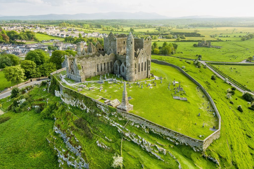 An aerial view of the Rock of Cashel in soft evening light. The roofless Gothic cathedral, the round tower and the fortified enclosure sit on a limestone outcrop above bright green fields, with a graveyard of Celtic crosses and headstones i
