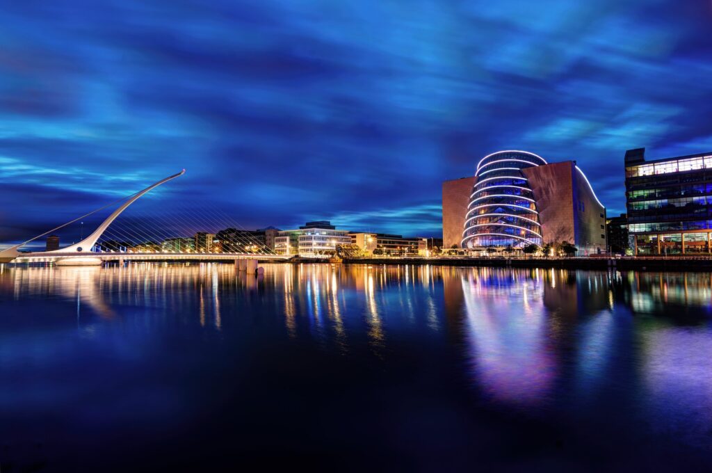 The harp-shaped Samuel Beckett Bridge and the illuminated tilted glass atrium of the Convention Centre Dublin glow at blue hour over the River Liffey in Dublin's Docklands, Ireland, their lights mirrored in the still, deep blue water beneat