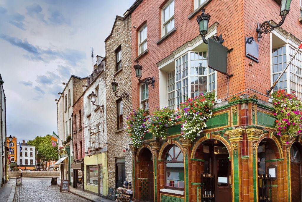 A cobbled street in the Temple Bar district of Dublin, Ireland. On the right corner stands a traditional pub with an ornate glazed-tile front in green, amber and gold, arched doorways and windows, wrought-iron lanterns and hanging baskets o